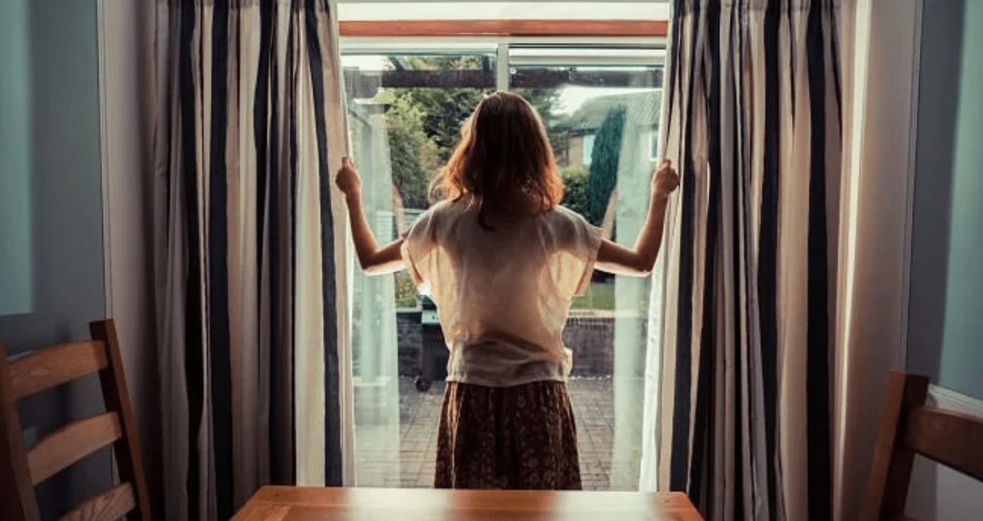 Young adult woman opening the curtains and looking out at her backyard