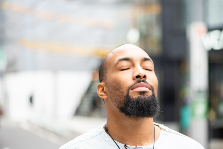 A handsome man standing outside with his eyes closed, enjoying a moment of peace.