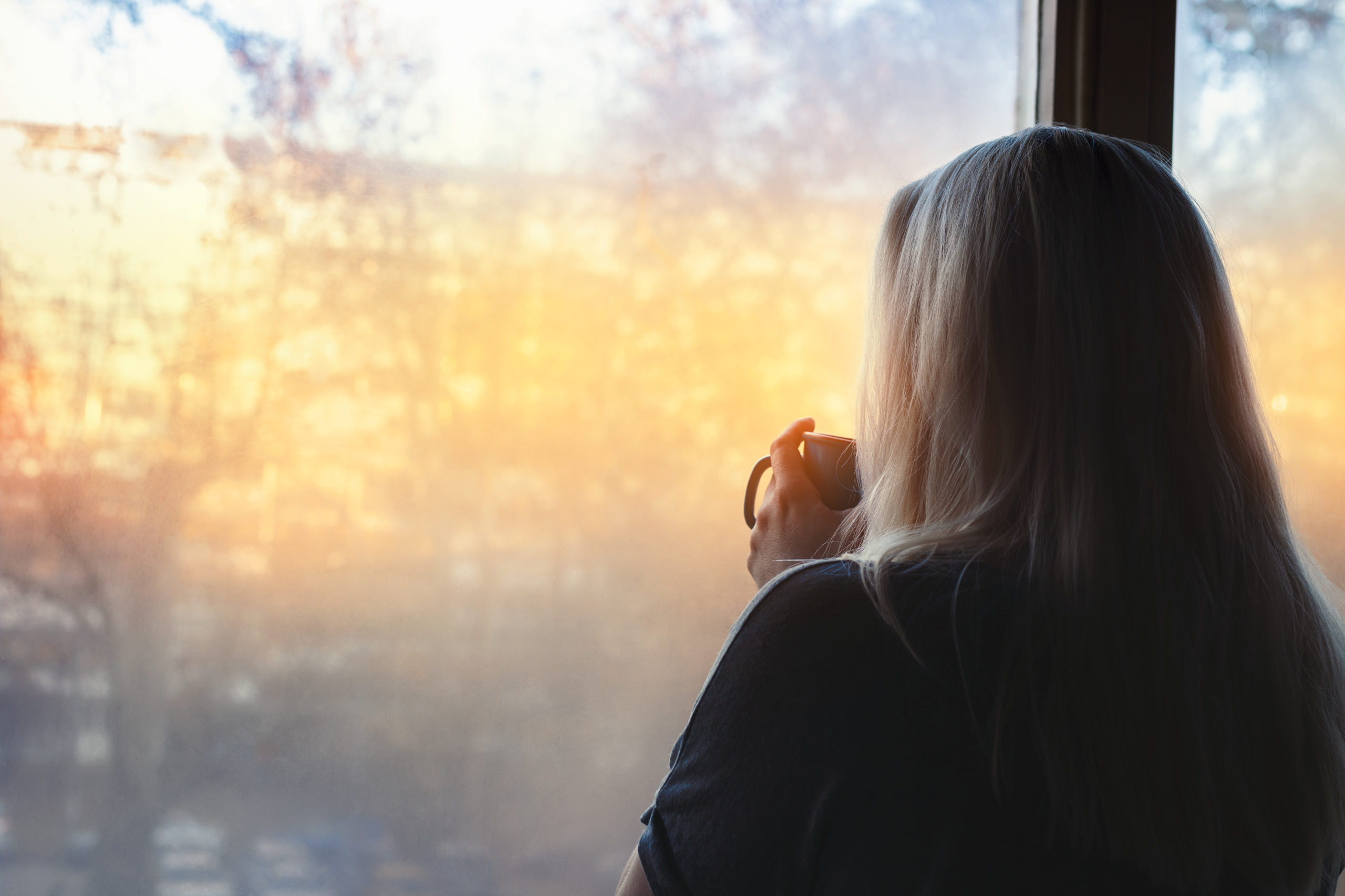 Blonde woman standing by the window, with coffee cup in hands, looking out into the morning light