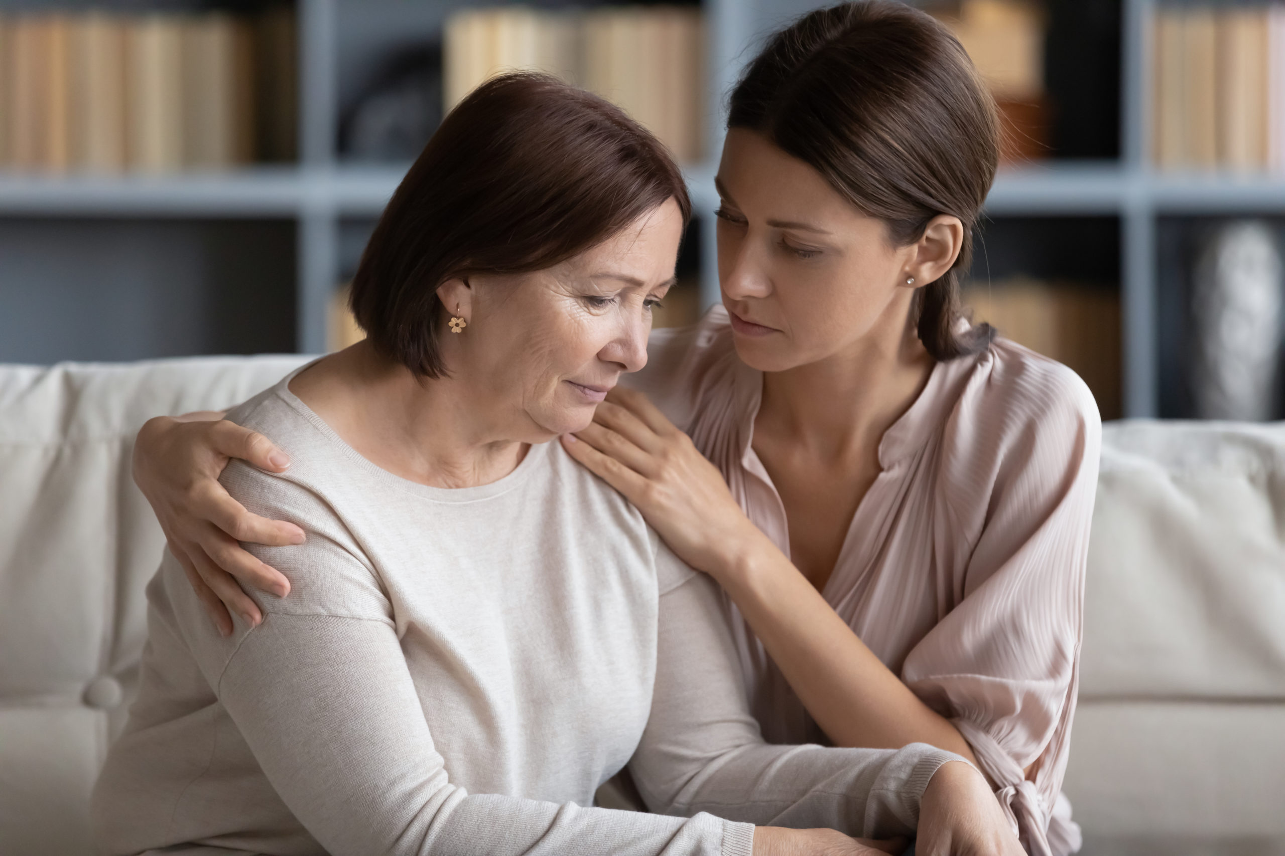 Young compassionate woman embracing shoulder of unhappy stressed middle aged mommy, comforting her at home. Kind grownup daughter supporting caring of older mature mother, sitting together on couch.