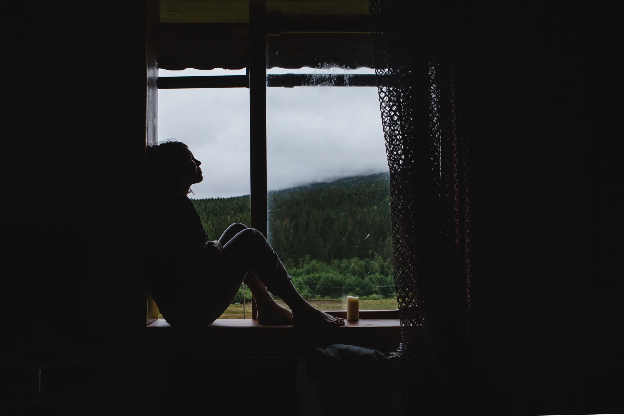 the girl is sitting on the windowsill. outside the window is a view of the mountains in the fog