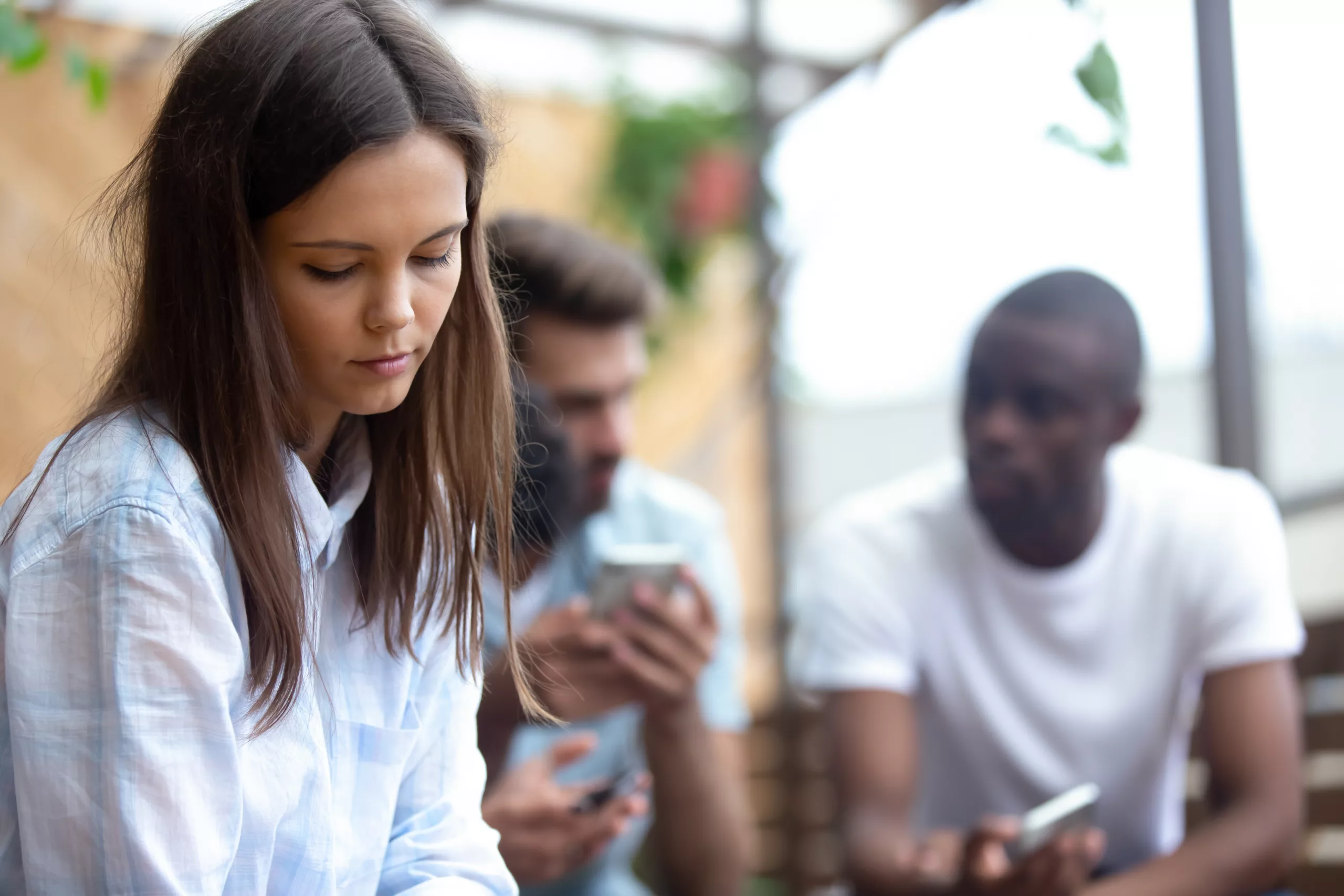 Multi-ethnic students spend time together gather in summer cafe talking using smartphones, close up focus on lonely frustrated pretty woman sitting separately from friends feels jealous or loneliness