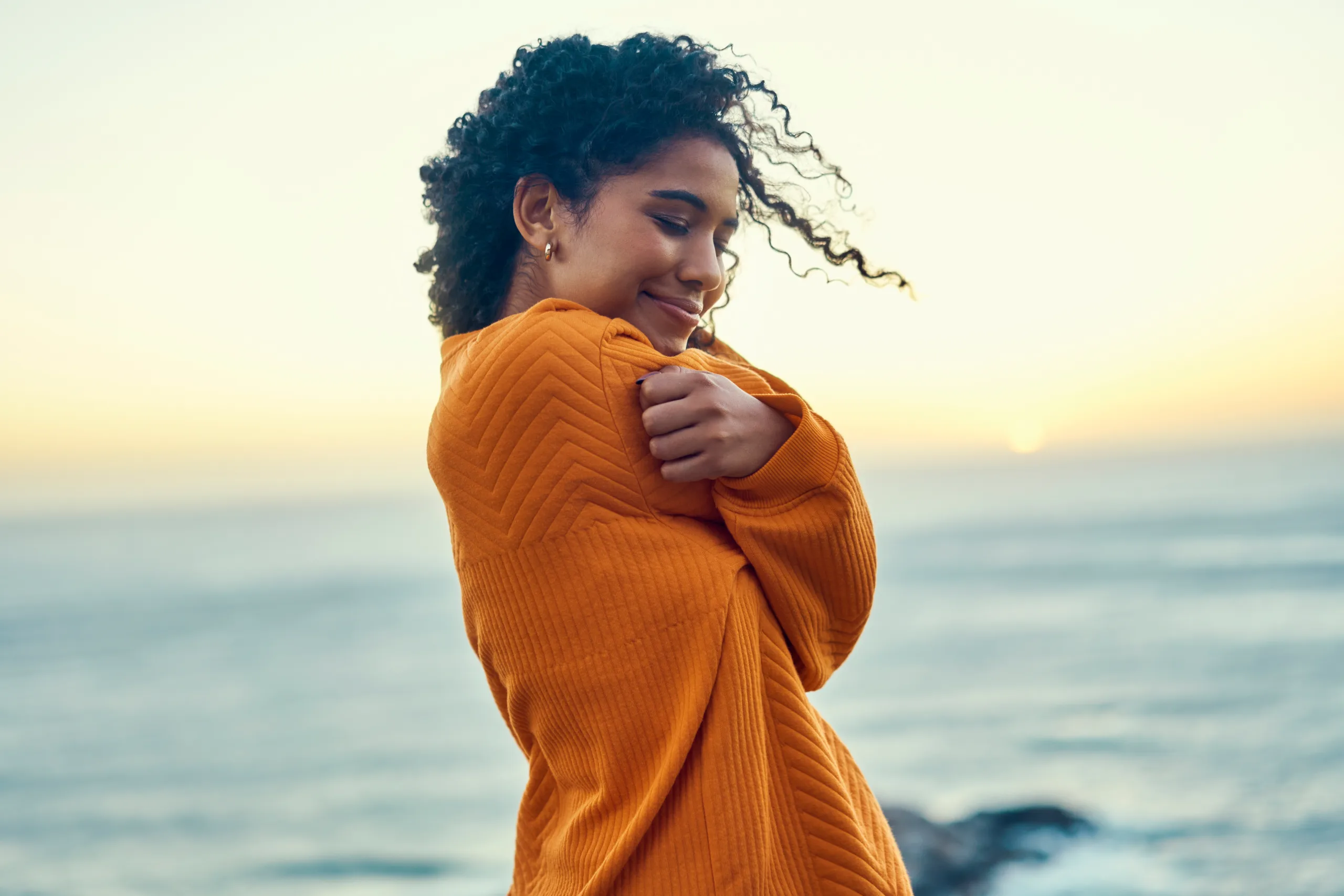 Happy black woman embrace herself on a beach at sunrise, self love and peaceful. Young African American feeling in touch with earth and nature, enjoy freedom and inner peace, smiling, loving soul